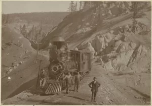 Three men stand next to a locomotive with their hands on their hips. One man sits on the front of the locomotive.