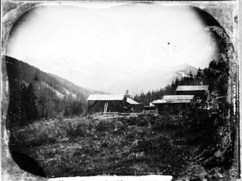 A small mining camp sits at the foot of the mountains. There is one building on the left side of the valley and two on buildings are on the right.