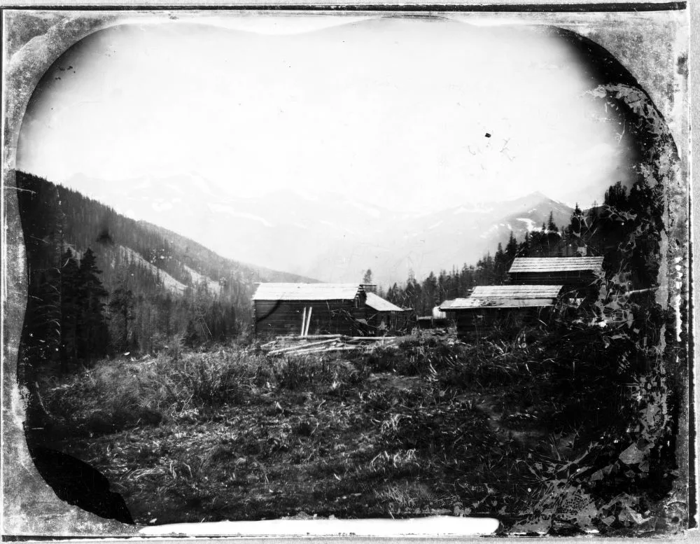 A small mining camp sits at the foot of the mountains. There is one building on the left side of the valley and two on buildings are on the right.