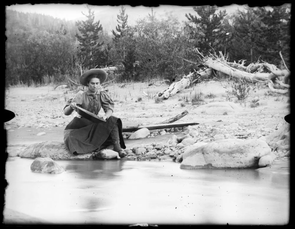 Mary Marks holds a gold pan and kneels at the edge of a river to pan for gold.