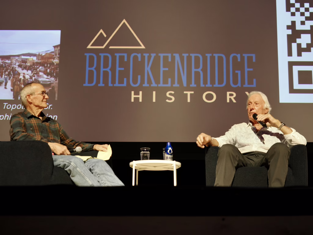 photo by Elaine Collins Cj Mueller and Lee Lucas sit on stage during the Breckenridge Rewind premier at the Riverwalk Center. A screen in the background shows the Breckenridge History logo.