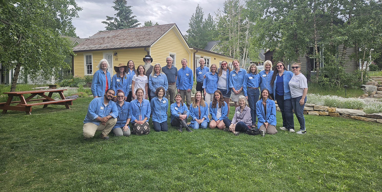 About 25 people in denim shirts kneel and stand in the grass to pose for a picture with a yellow house in the background.