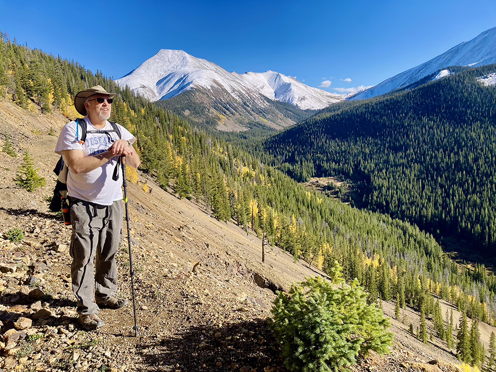A man in a white shirt and grey pants leans on a hiking pole and gazes off to the right with snow-covered mountain peaks behind him.